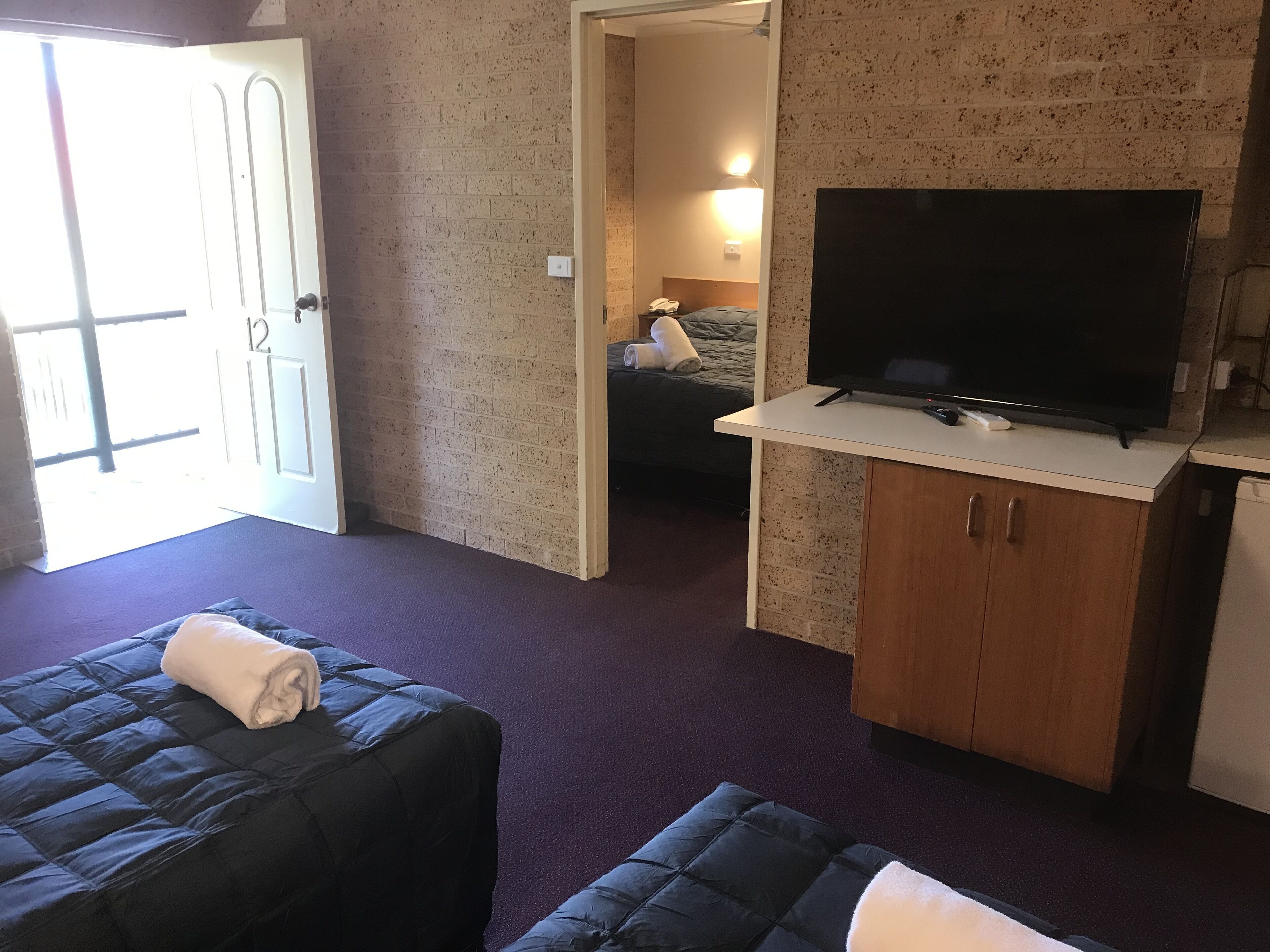 Habitación de hotel con cama, televisor sobre un pedestal, armario de madera y puerta que da a otra habitación.