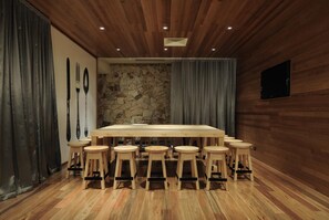 Une salle de conférence dotée d’une grande table en bois entourée de tabourets, d’un mur en pierre, de lambris en bois et d’un téléviseur fixé au mur.