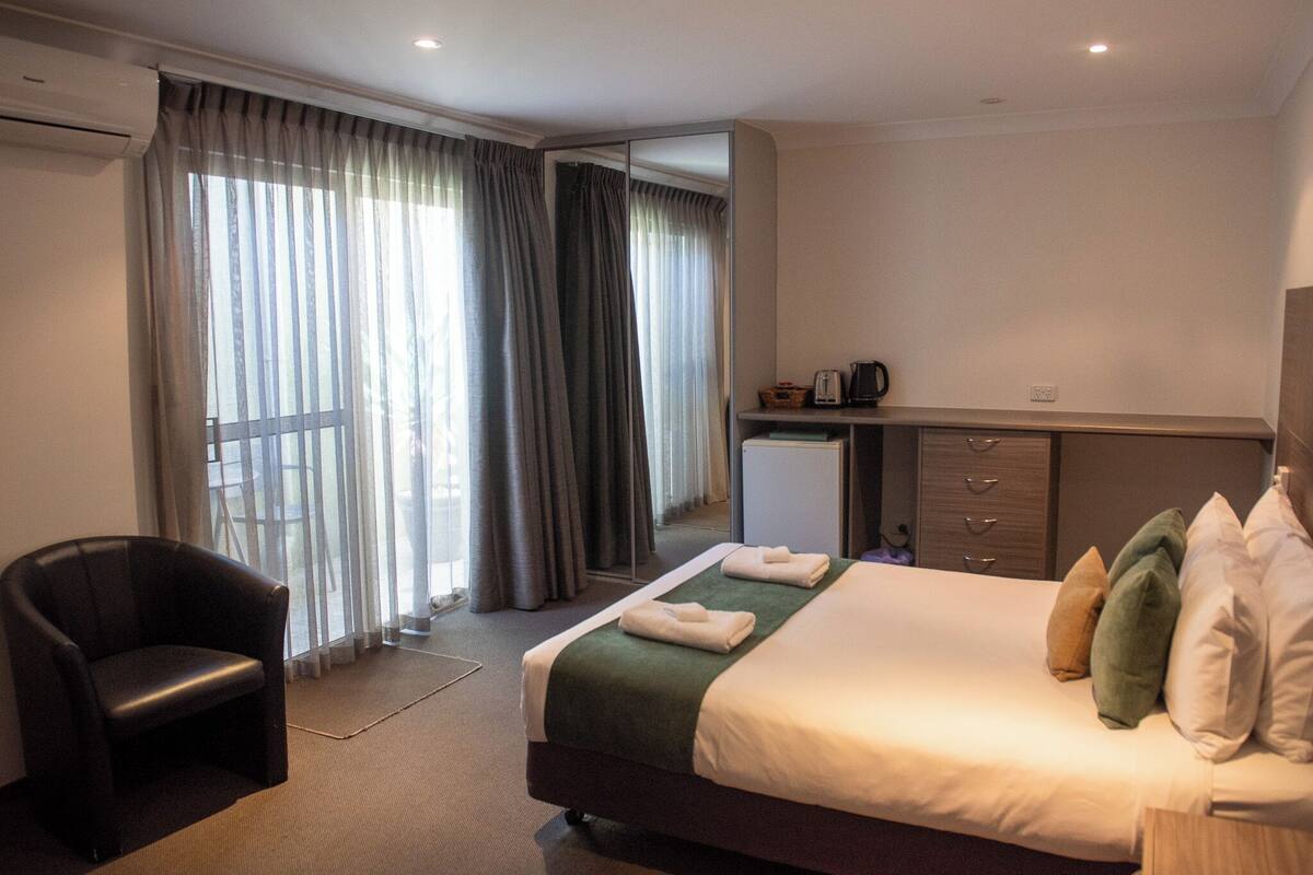 A hotel room with a bed, a chair, a desk with a computer, and a window with curtains.