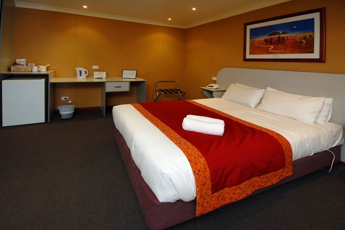 A hotel room with a bed, a desk, a chair, a small table, a nightstand, and a painting on the wall.