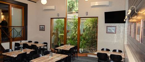 A restaurant with multiple tables and chairs, large windows with a view of greenery, and an air conditioning unit on the wall.