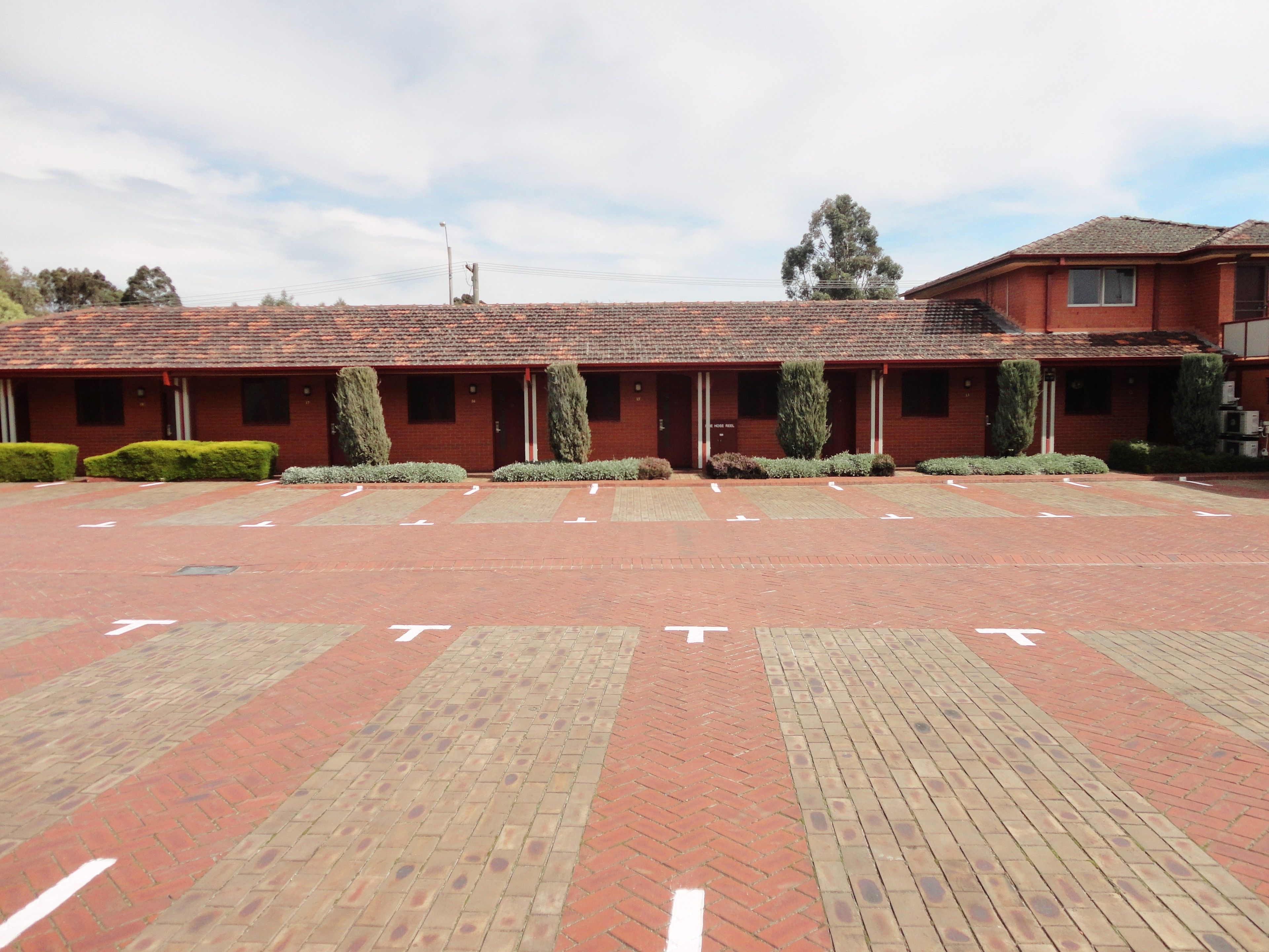 A red-brick building with a tiled roof, a parking lot with directional arrows, and neatly trimmed hedges.