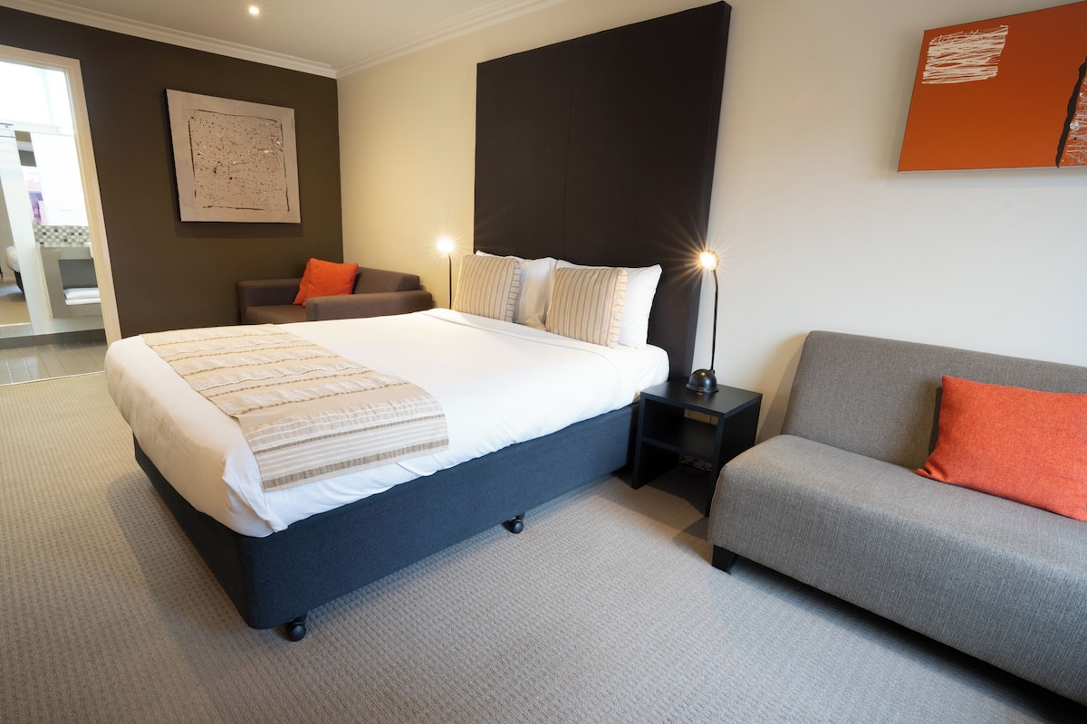 A modern bedroom with a large bed, a grey sofa, and a nightstand with a lamp.