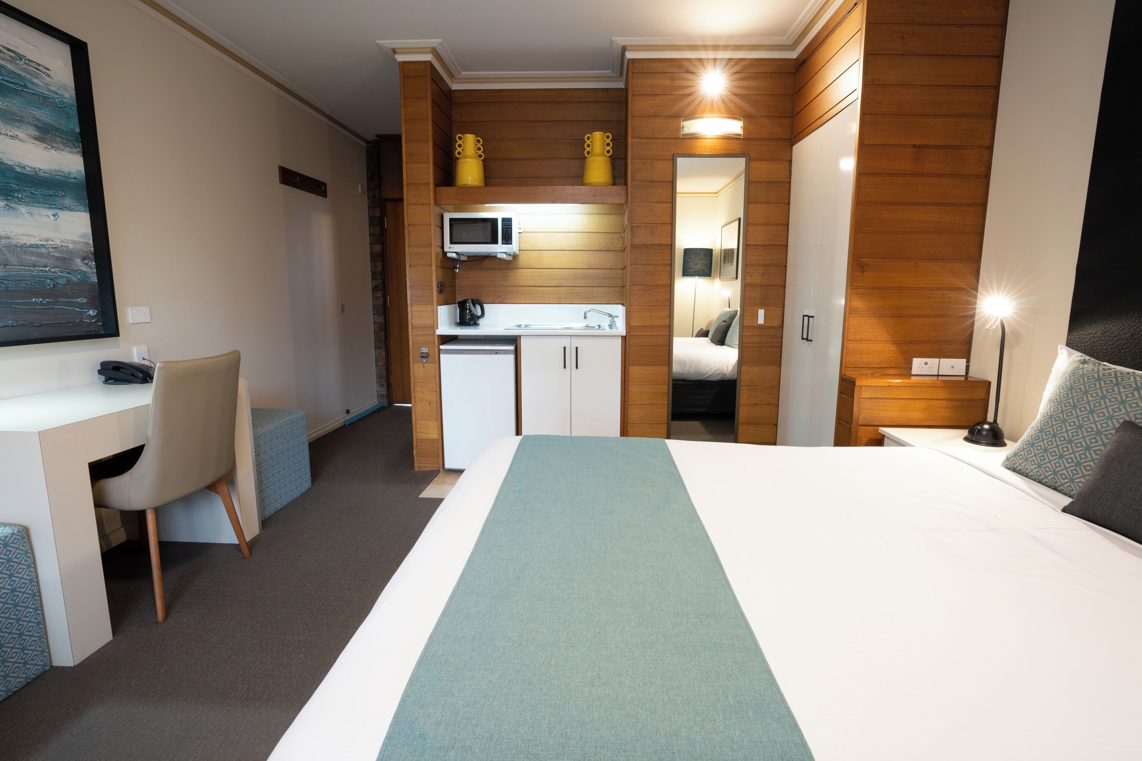 Ett kompakt hotellrum med en säng, ett skrivbord, ett litet kök och ett badrum.