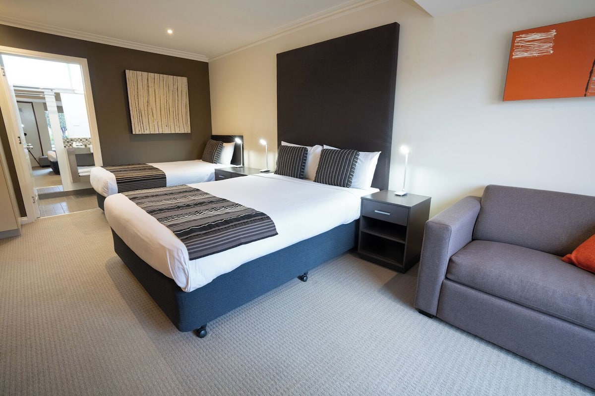 A hotel room with a large bed, a sofa, a nightstand, and a wall-mounted artwork.
