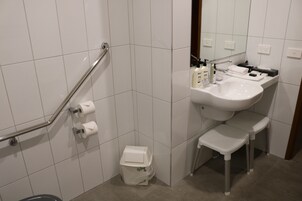 Casa de banho com azulejos brancos, pia, espelho, sanita com barra de apoio e um contentor de lixo.