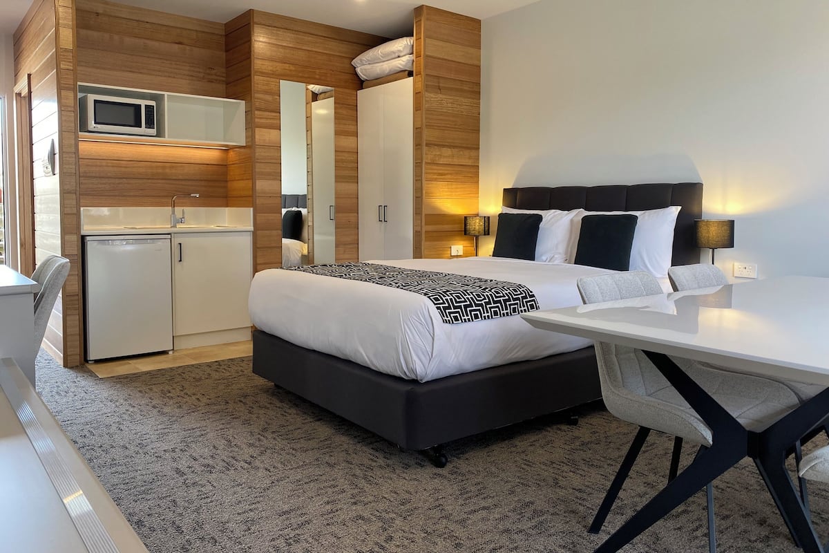 A modern hotel room with a bed, bedside table, desk, and a kitchenette area.
