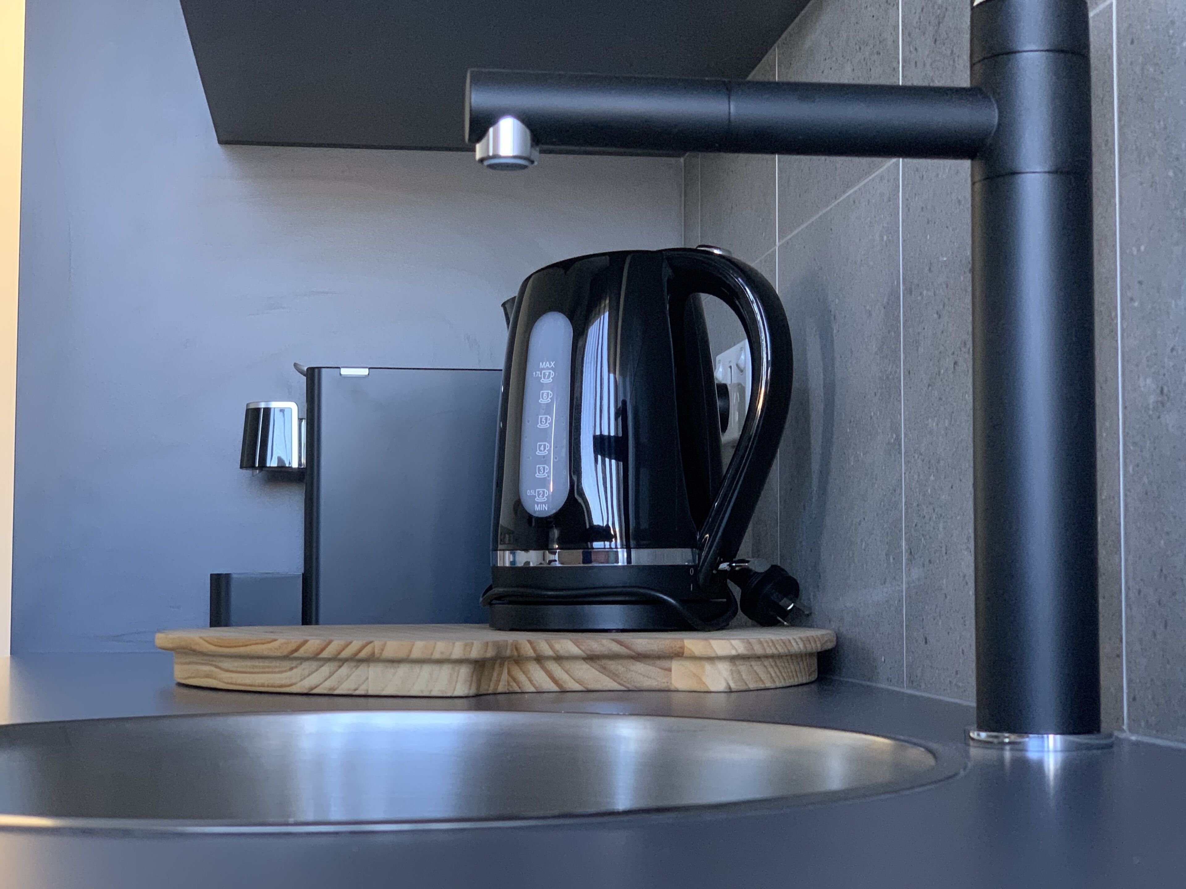 Una cocina moderna con un lavamanos de acero inoxidable, una tetera negra sobre una bandeja de madera y un grifo de pared.