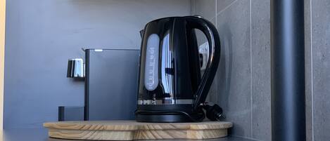 Cozinha moderna com uma pia de aço inoxidável, uma chaleira preta sobre uma bandeja de madeira e uma torneira fixa na parede.