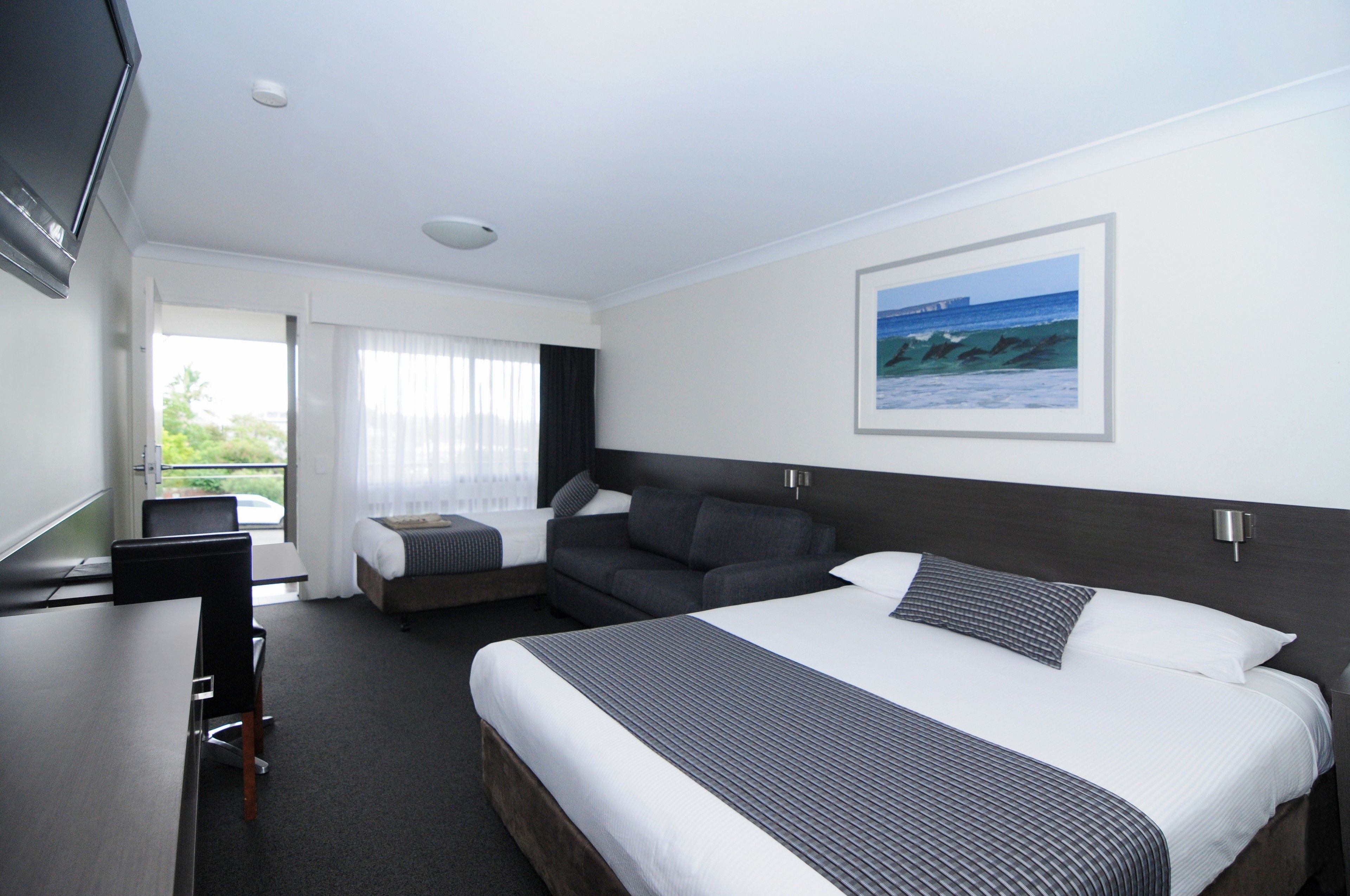 A hotel room with a large bed, a desk, and a chair. There is a window with curtains, a painting on the wall, and a view of the ocean.