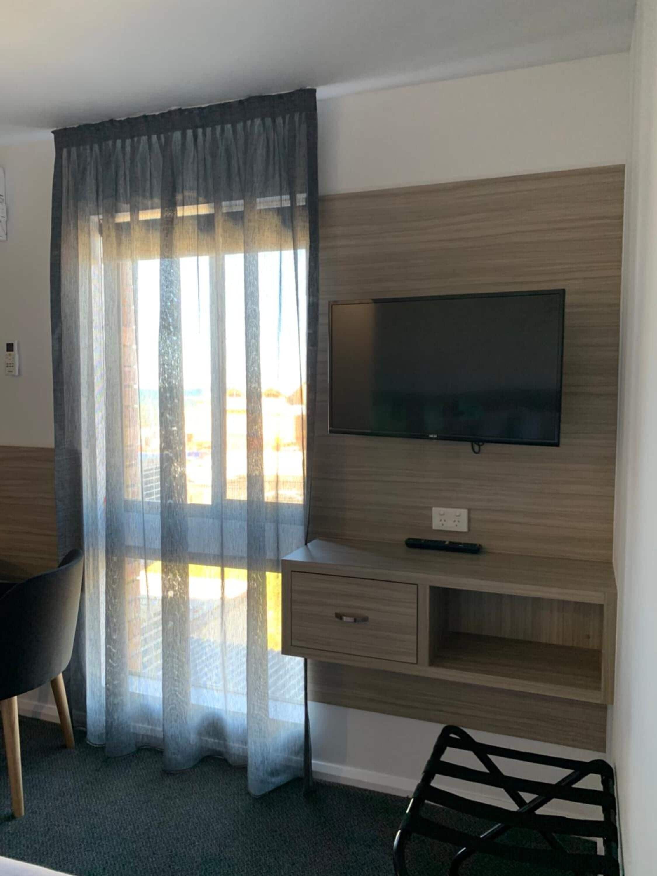 Une chambre d’hôtel avec un bureau en bois, une chaise, un téléviseur fixé au mur et une fenêtre ornée de rideaux légers.