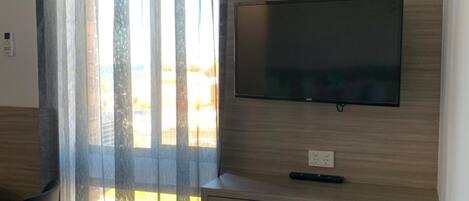 Ein Hotelzimmer mit Holztisch, einem Sessel, einem an der Wand montierten Fernseher und einem Fenster mit durchsichtigen Vorhängen.