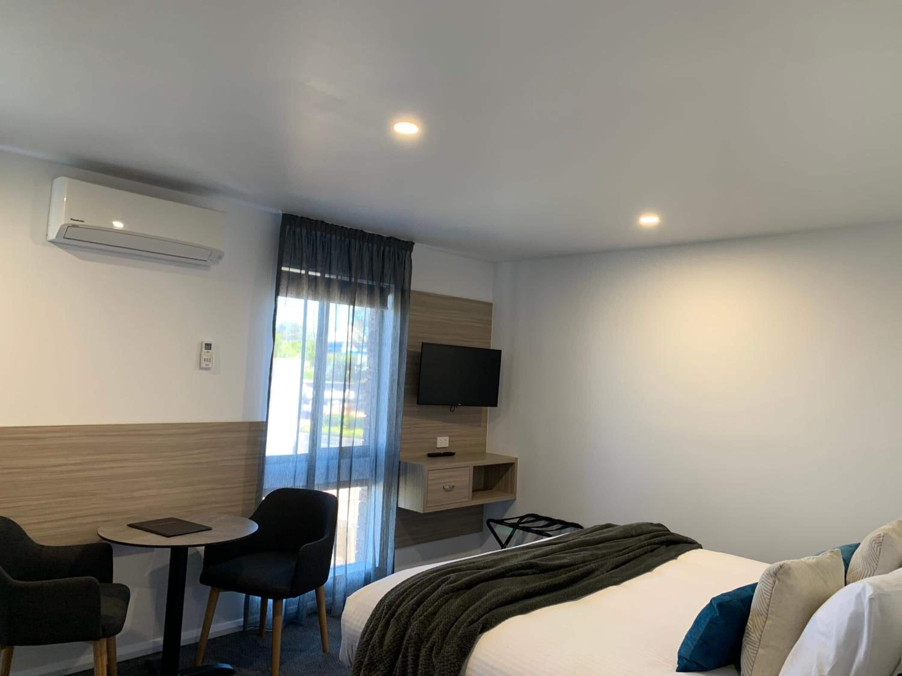 Una habitación de hotel con cama, escritorio con silla, televisor y aire acondicionado.