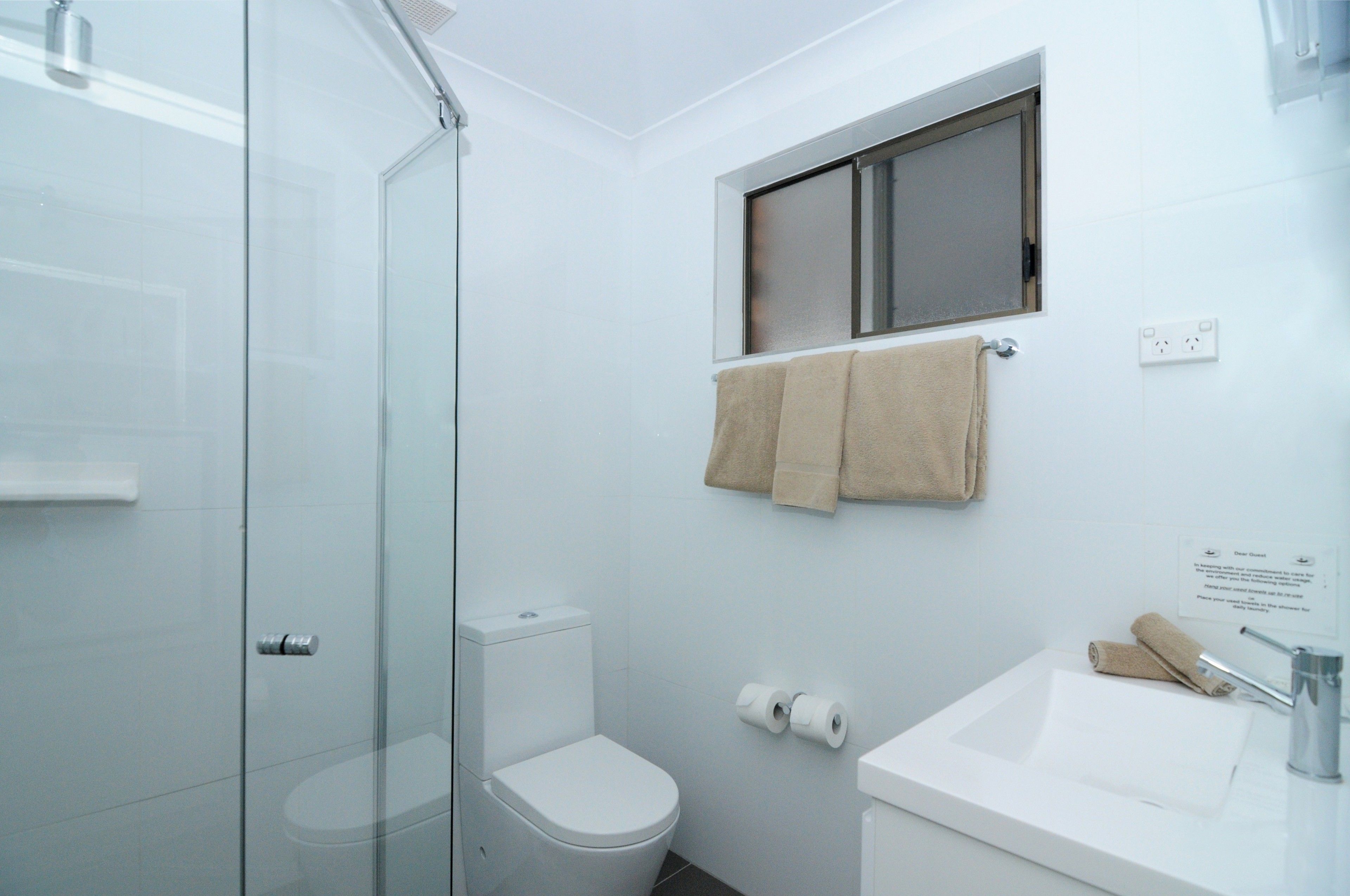 A modern bathroom with a glass shower enclosure, a toilet, a sink, and a window with a towel hanging on the wall.