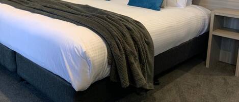 En pænt redt seng med et gråt tæppe og blå puder, et natbord med en lampe, og et tæppe på gulvet.