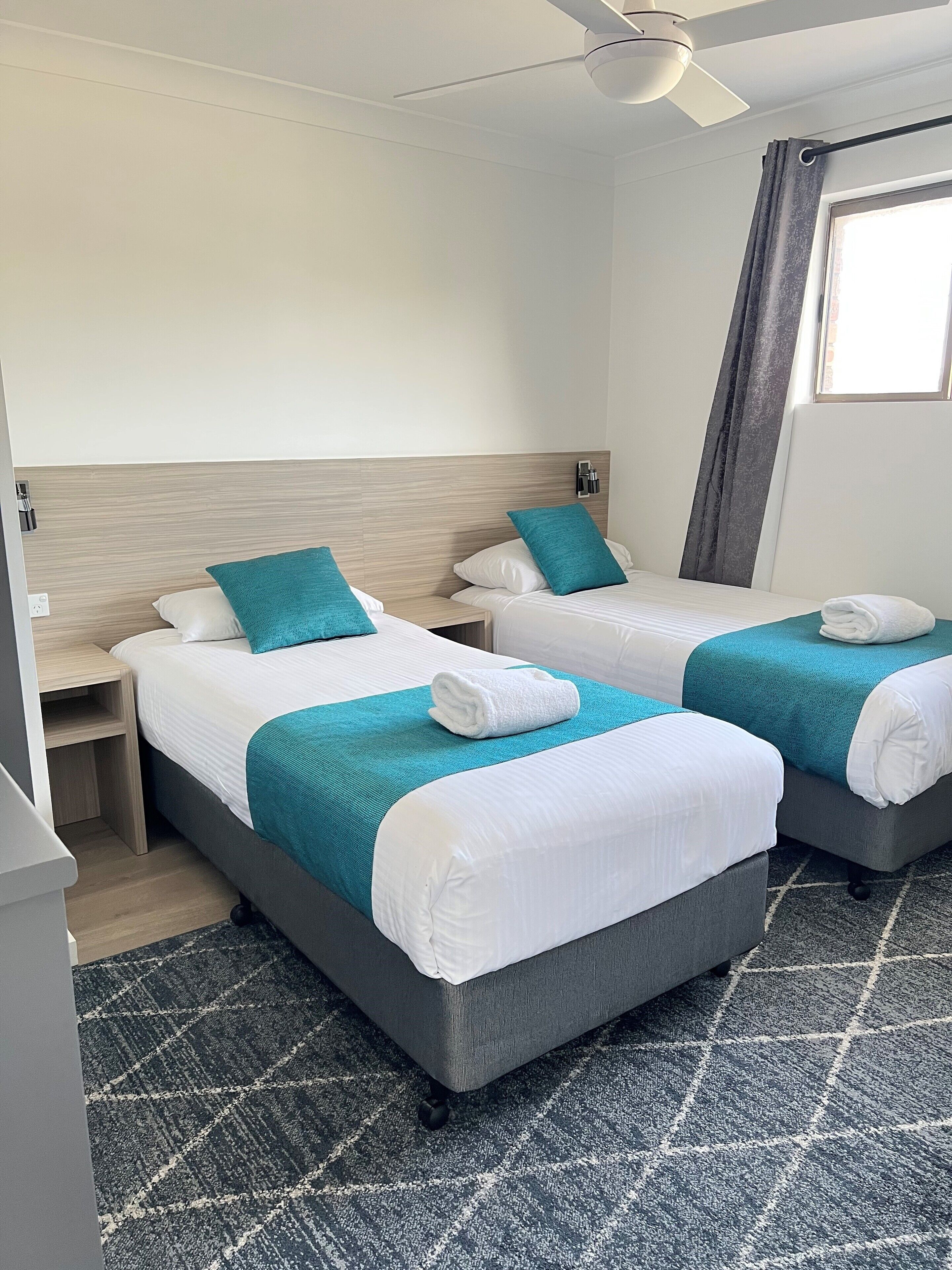 Habitación de hotel con dos camas, cada una con un edredón color turquesa y almohadas blancas, una mesita de noche pequeña, un ventilador de techo y una ventana con cortinas.