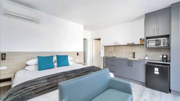 A modern hotel room with a bed, a blue armchair, a kitchenette with a microwave, and a refrigerator.