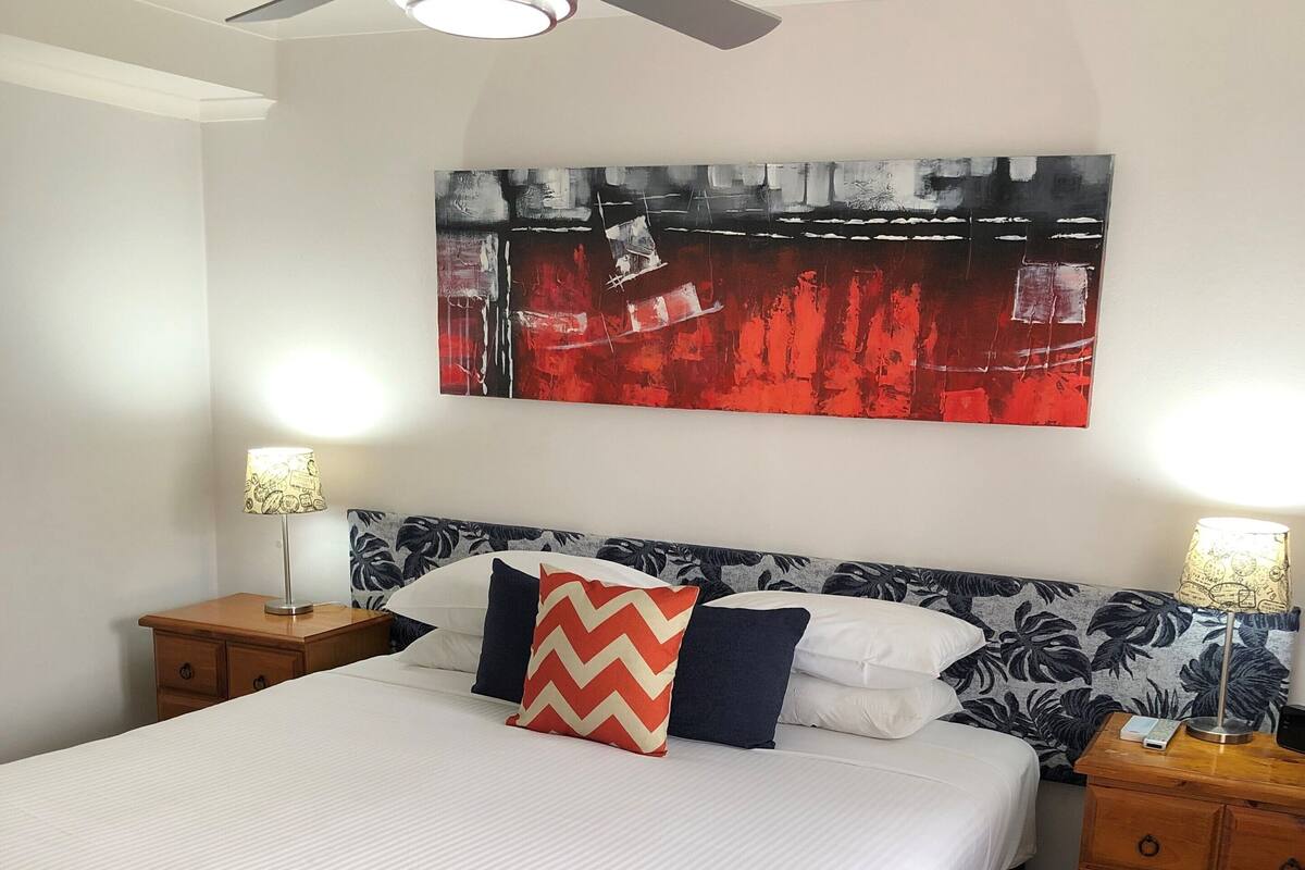 A neatly made bed with a white and navy blue duvet, two nightstands with lamps, a ceiling fan, and a large abstract painting on the wall.