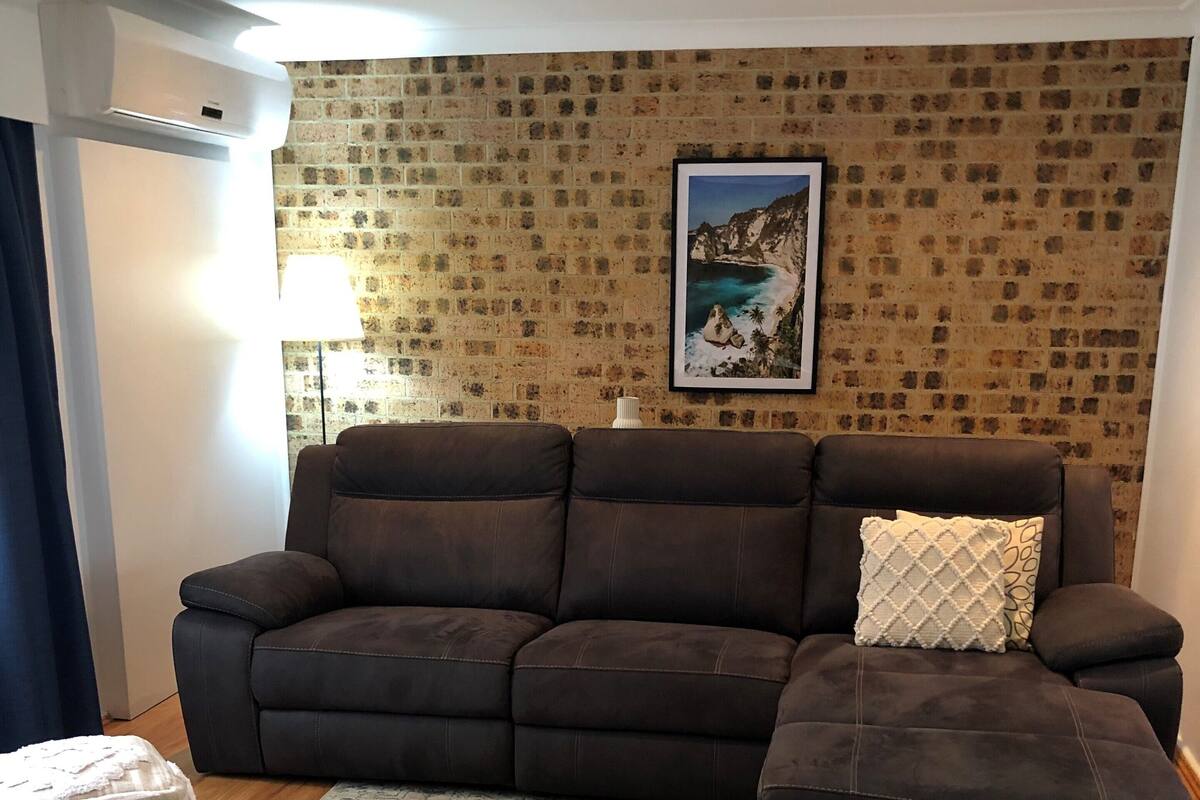 A living room with a brown sectional sofa, a patterned rug, and a brick-patterned wall.