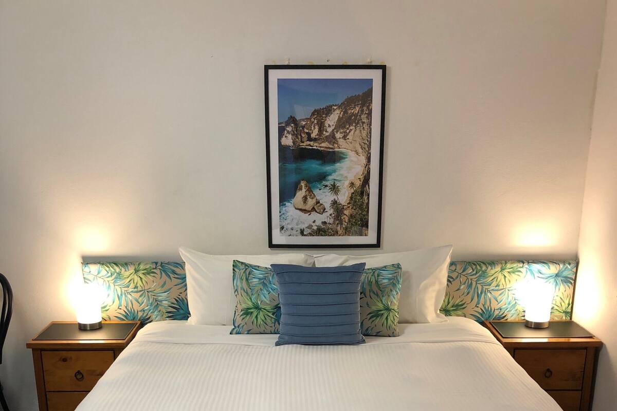 A neatly made bed with a white duvet, flanked by two nightstands with lamps, a framed picture of a coastal scene, and a pillow with a tropical leaf pattern.