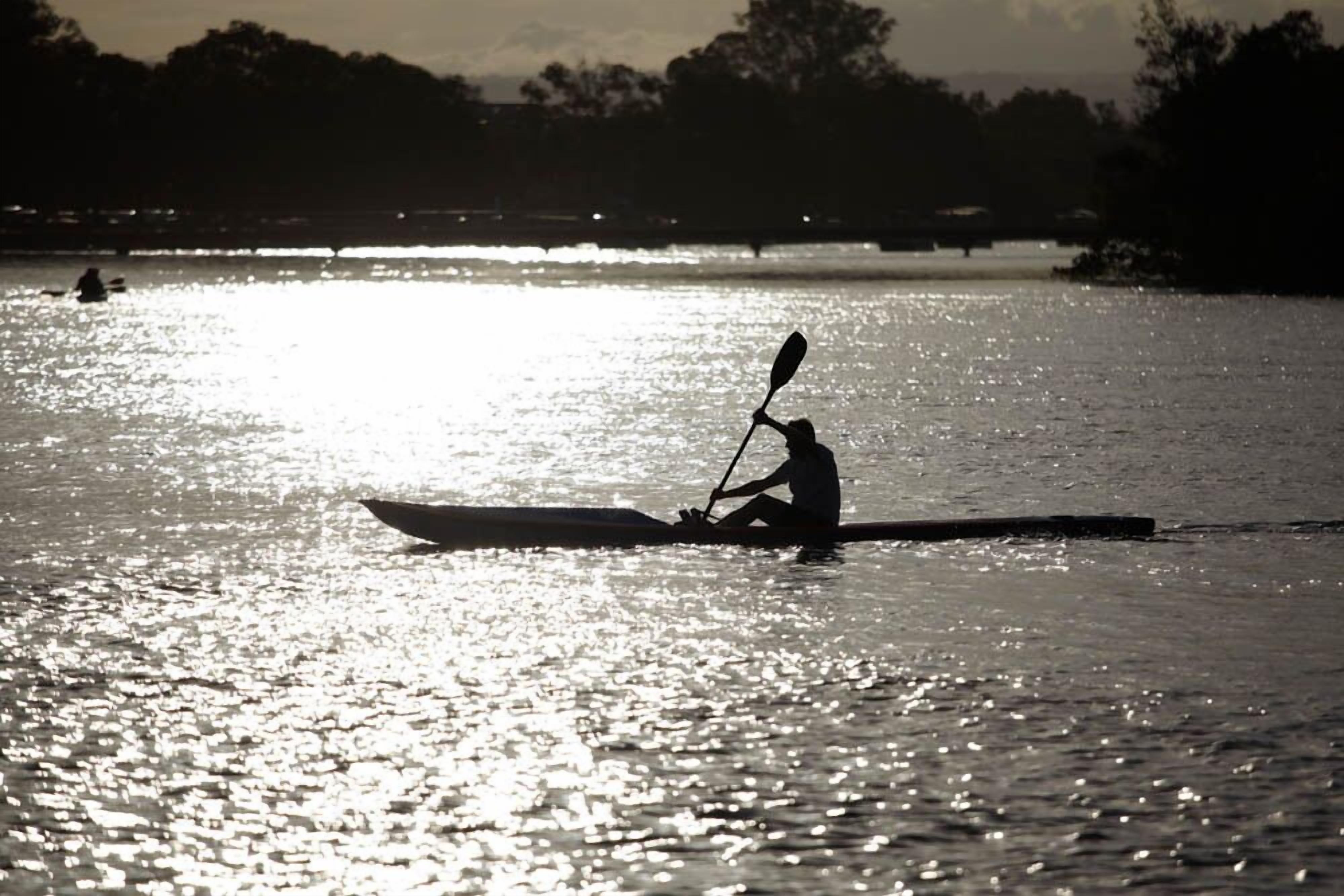 A silhouette of a person kayaking on a calm body of water during sunset.