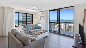 Un salon moderne avec un canapé, une table basse et un téléviseur. Il est doté de grandes fenêtres offrant une vue sur la plage et l’océan.