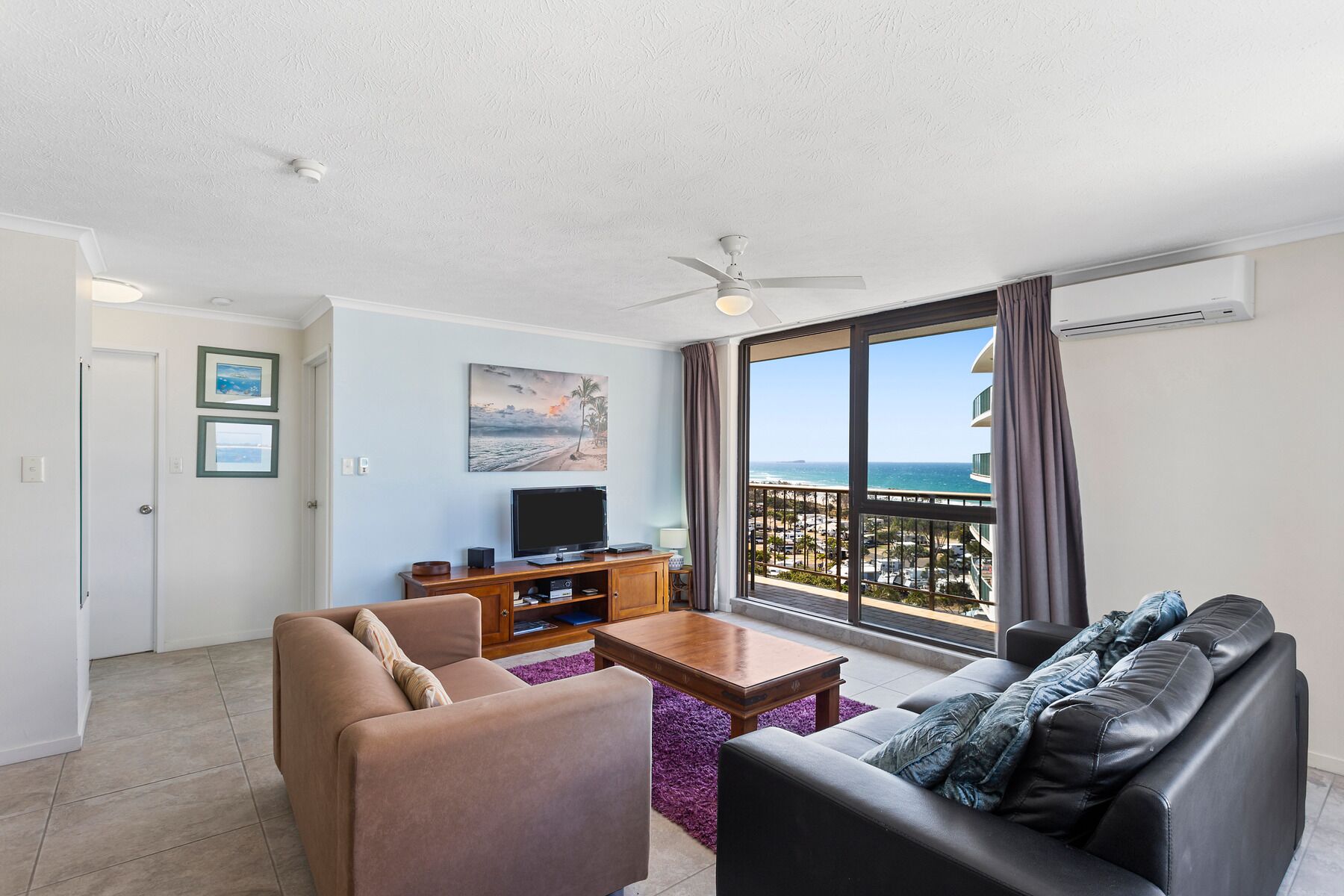 A living room with a TV, sofa, and coffee table, a balcony with a view of the ocean, and a ceiling fan.