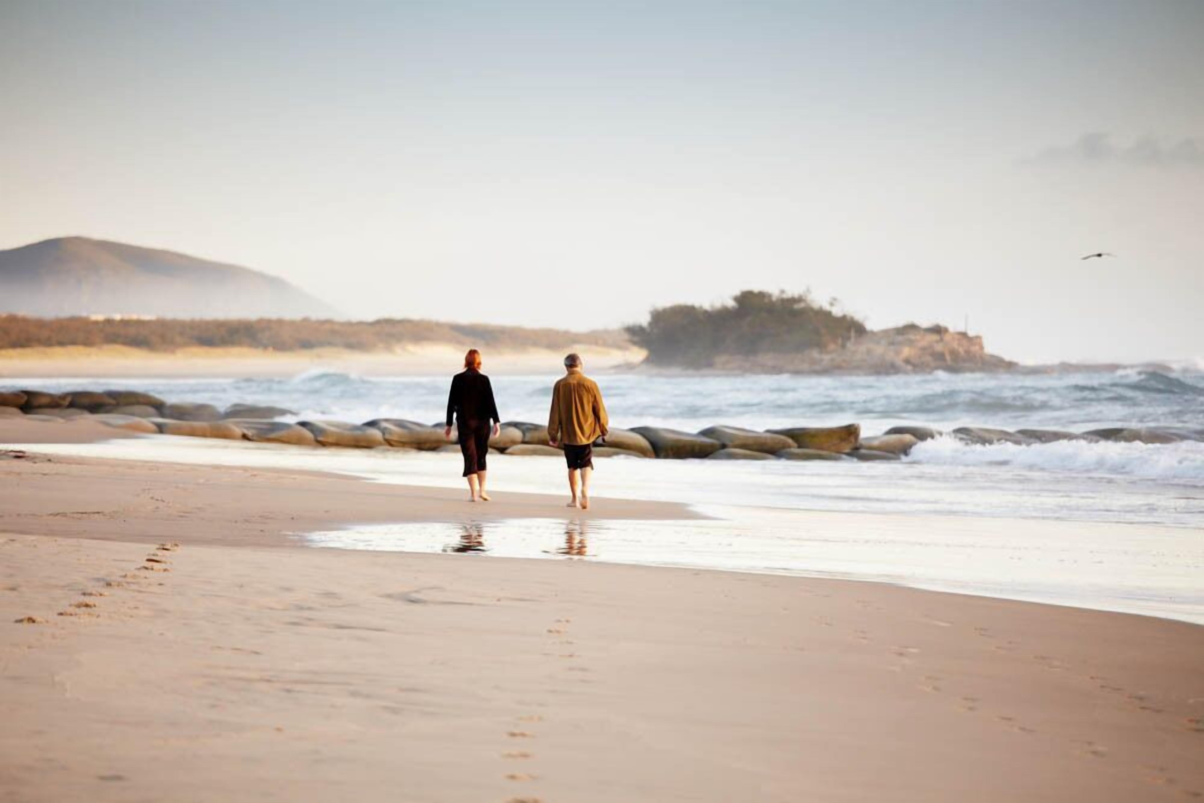 Two people walking on a beach with waves and a rocky outcrop in the background.