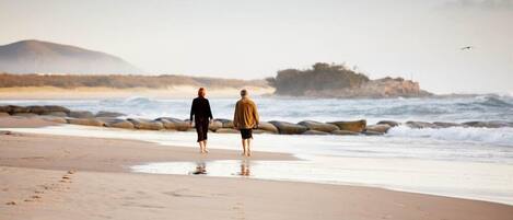 Two people walking on a beach with waves and a rocky outcrop in the background.