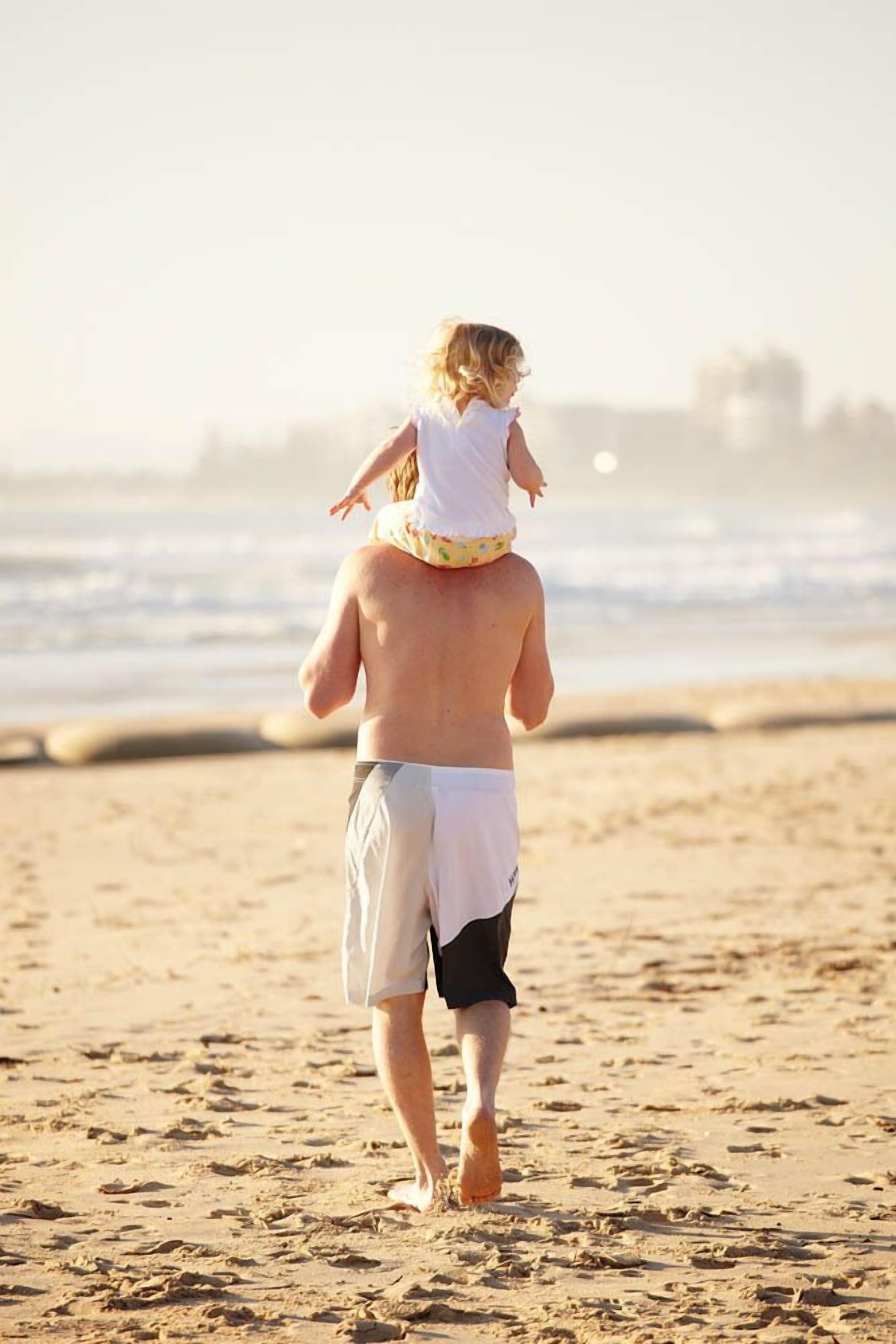 A person carrying a child on their back on a sandy beach with the ocean in the background.