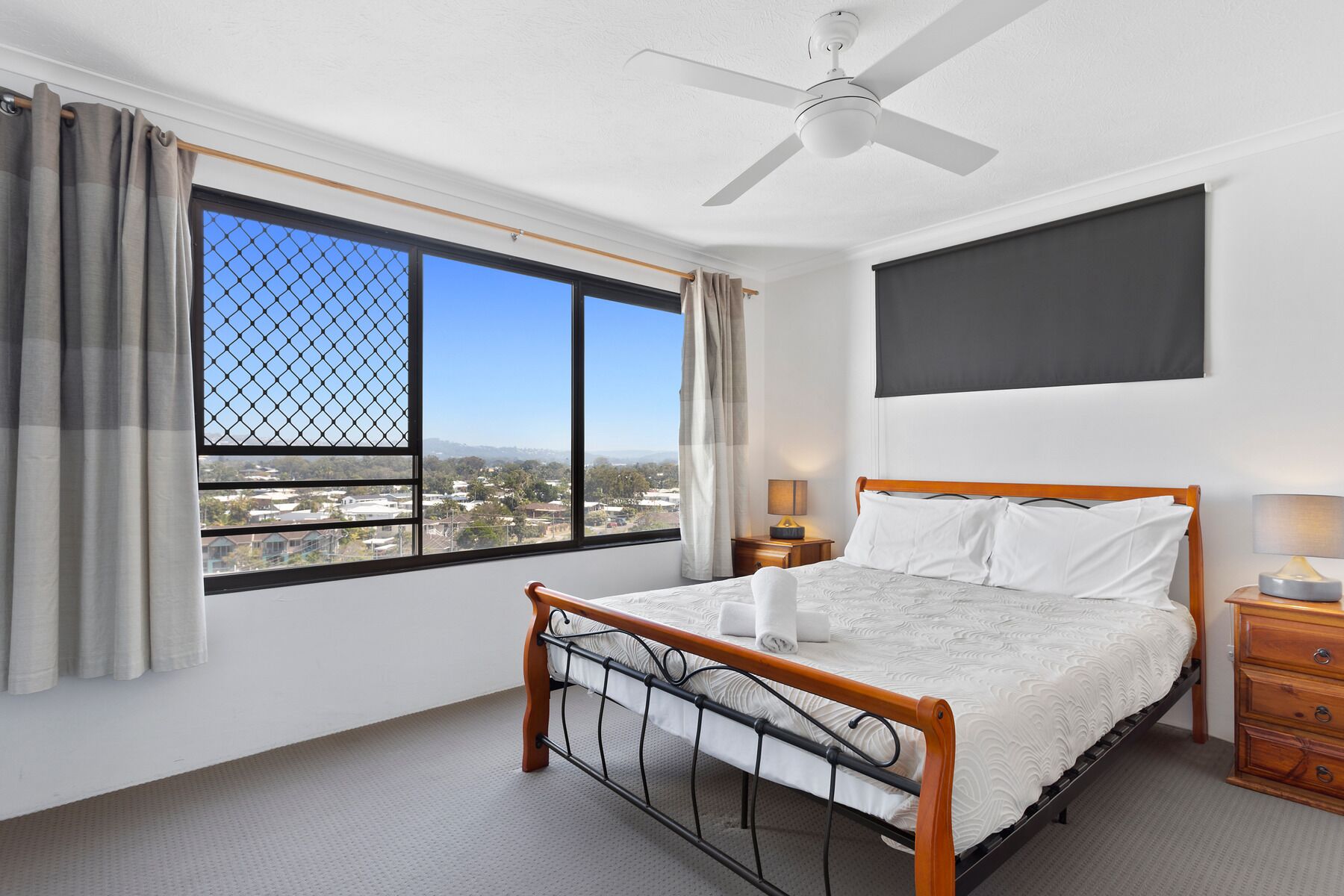 A bedroom with a bed, a ceiling fan, a window with curtains, and a view of the city.