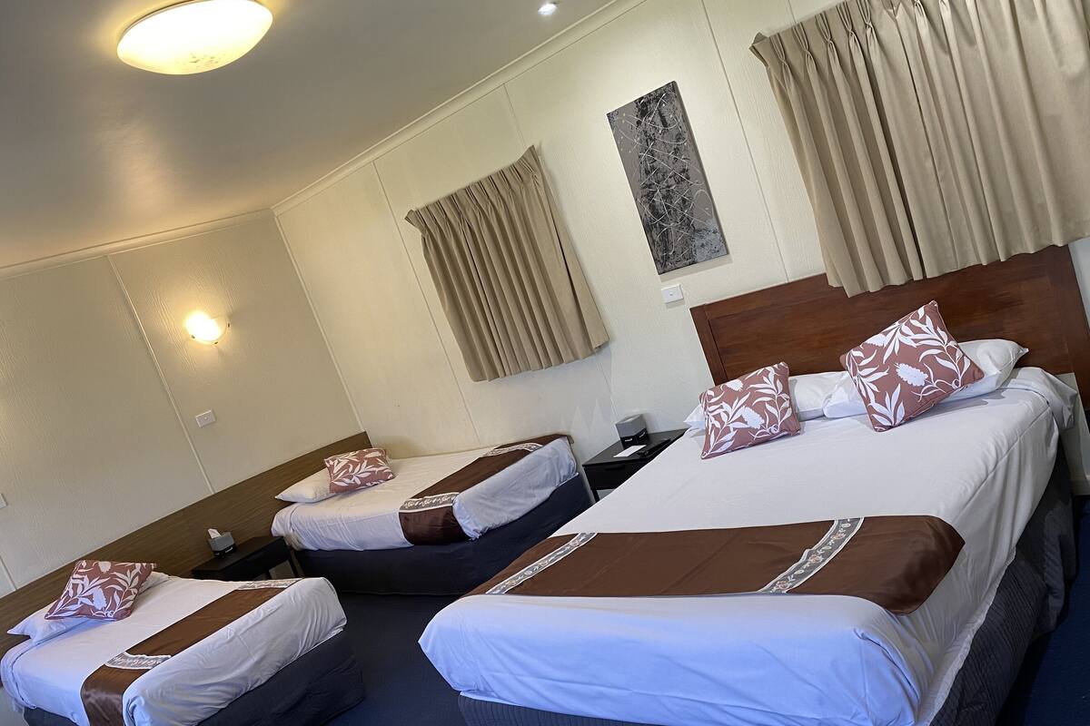 Habitación de hotel con tres camas, cada una con almohada de diseño marrón y blanco, cabecera de madera y mesita de noche con lámpara.