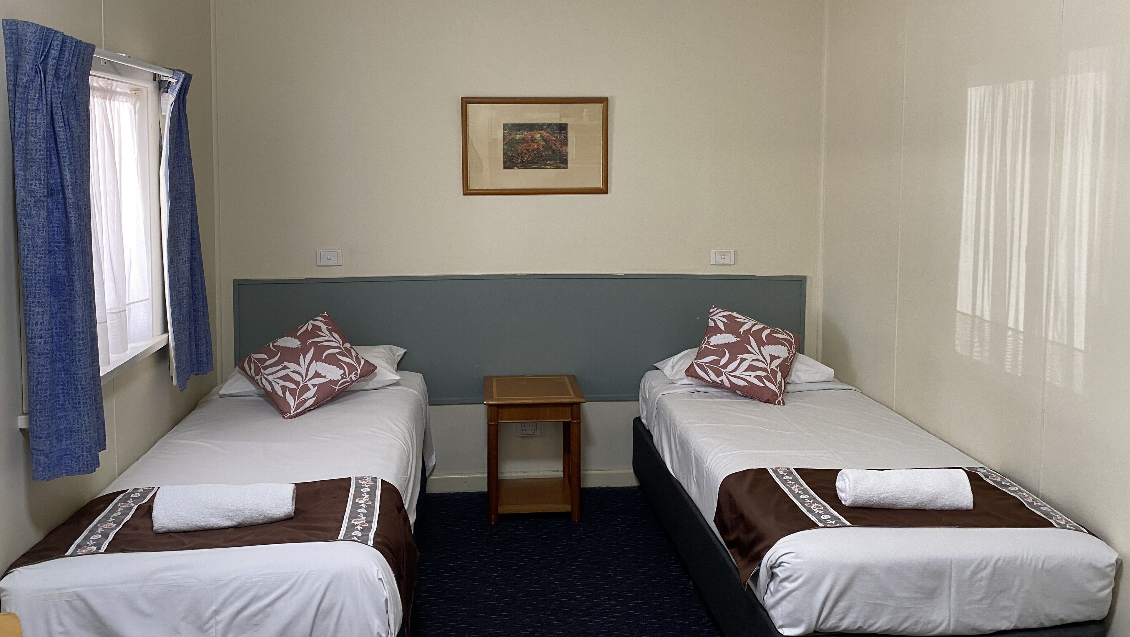 A room with two single beds, a small wooden nightstand, a framed picture on the wall, and a window with blue curtains.