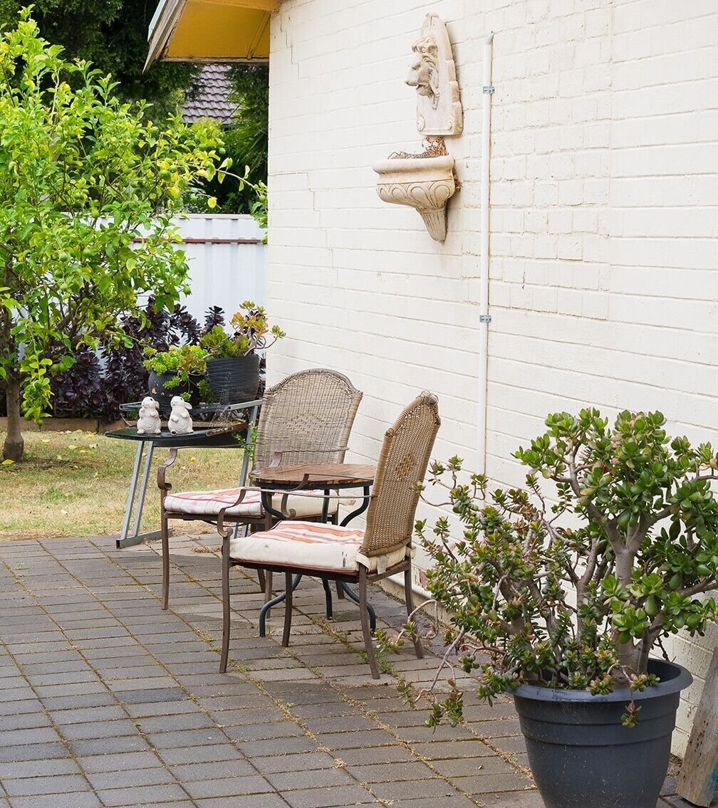 A patio with two wicker chairs and a small table, a potted plant, and a decorative stone feature on a white wall.