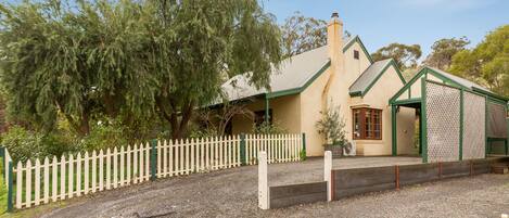 A house with a gravel driveway, a white picket fence, and a large tree in the background.