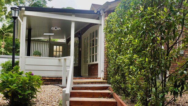 A covered outdoor area with steps leading to a door, surrounded by greenery and a brick wall. - Curtis Falls Apartment (Tamborine Mountain)