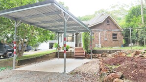 A covered outdoor area with a brick building in the background, a concrete pathway, and a parked vehicle. - Curtis Falls Apartment (Tamborine Mountain)
