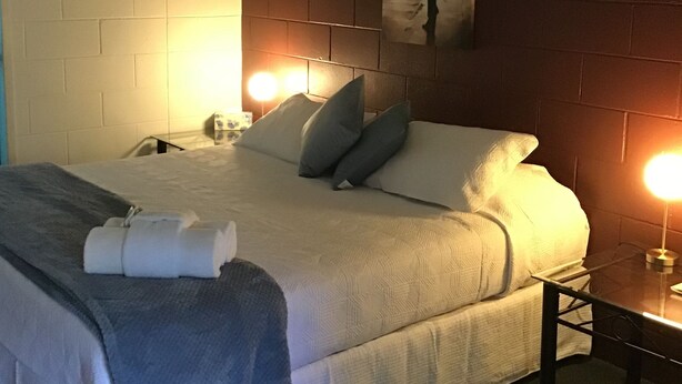 Een hotelkamer met een bed, kussens, een nachtkastje, een lamp en een wandairconditioner.