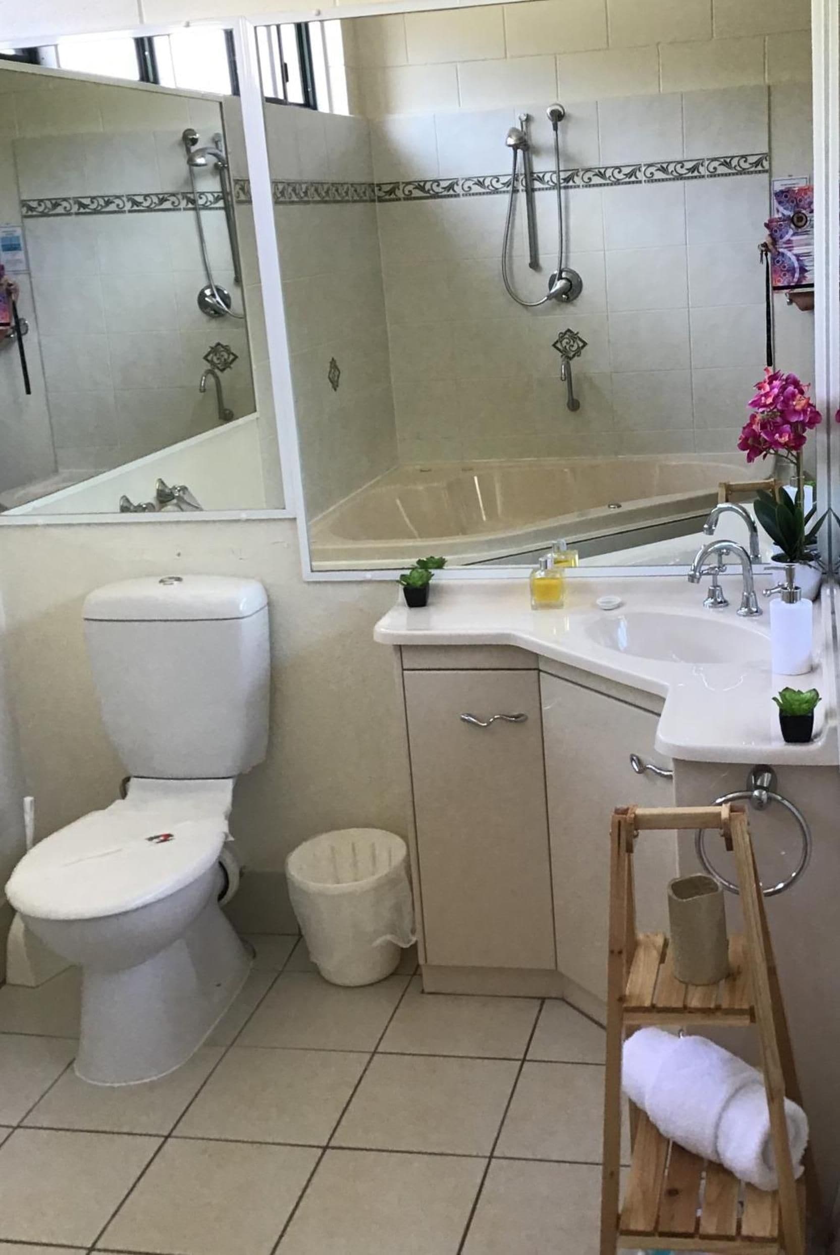 Un baño con inodoro, lavamanos, espejo, ducha y un taburete de madera pequeño con una toalla.
