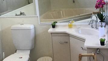 A bathroom with a toilet, sink, mirror, shower, and a small wooden stool with a towel.