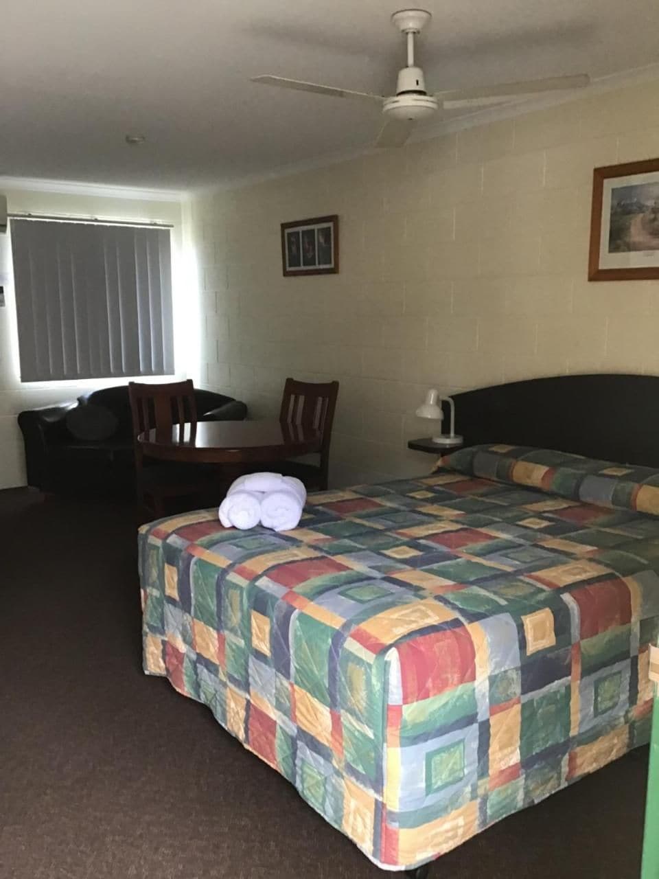 Ett hotellrum med en säng, ett skrivbord, en stol, en takfläkt och ett fönster med persienner.