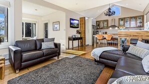 A modern living room with a dark leather sofa, a large rug, and a kitchen area with wooden cabinets and a dining set. - Topiary Haven (West Launceston)