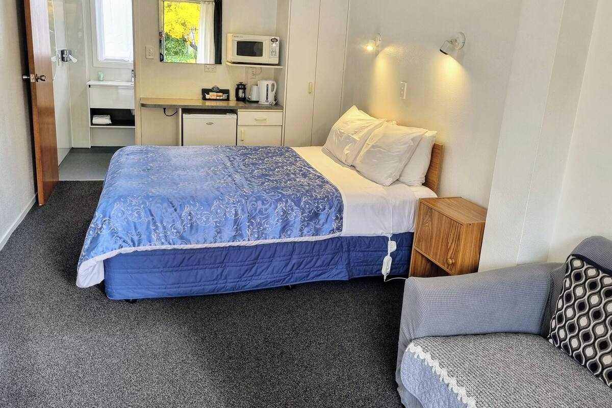 A bedroom with a bed, bedside table, and a small seating area. There is a kitchenette with a microwave, kettle, and coffee maker.