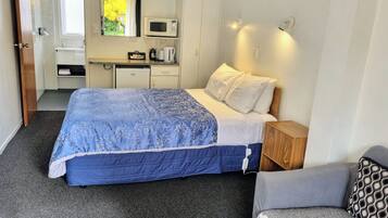 A bedroom with a bed, bedside table, and a small seating area. There is a kitchenette with a microwave, kettle, and coffee maker.