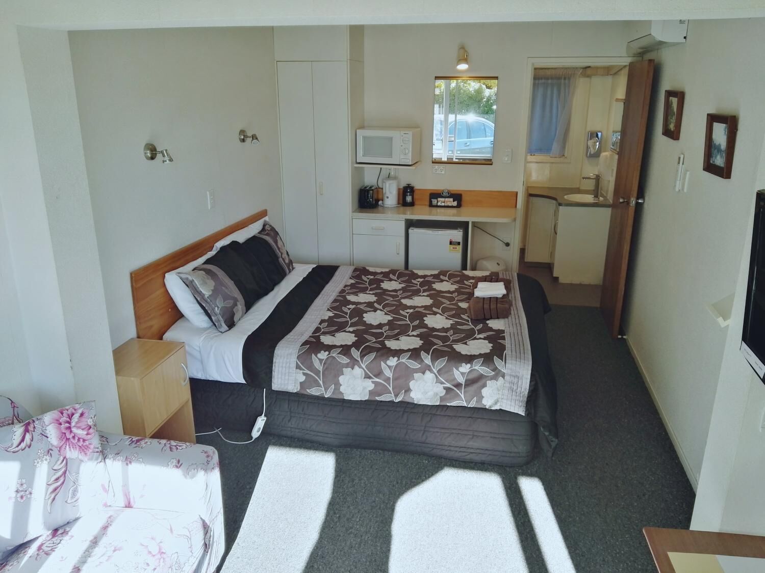 A compact hotel room with a bed, bedside table, a small desk with a chair, a microwave, a refrigerator, and a window with a view of the outside.