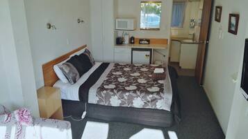 A compact hotel room with a bed, bedside table, a small desk with a chair, a microwave, a refrigerator, and a window with a view of the outside.