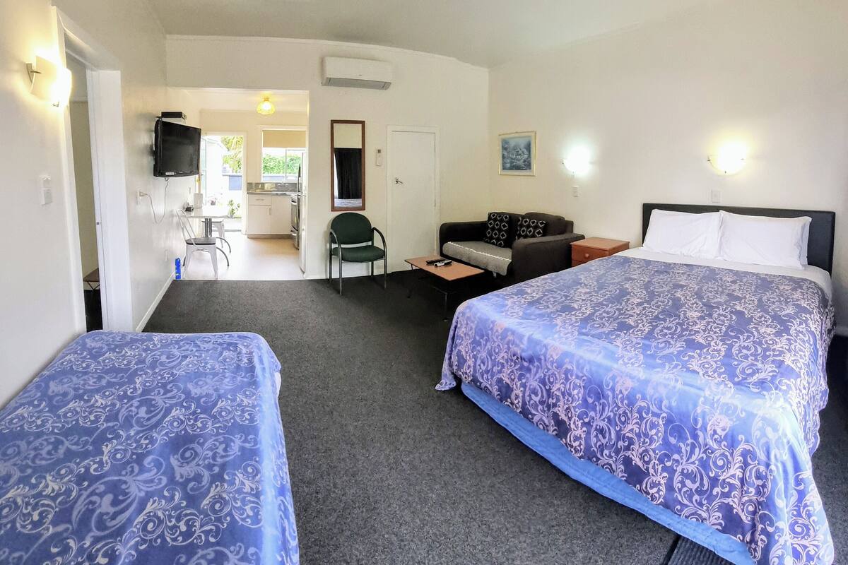 A hotel room with two beds, a sofa, a small table, and a kitchenette in the background.