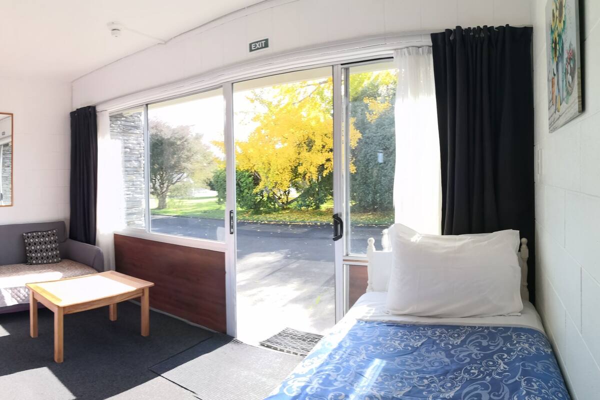 A hotel room with two beds, a sofa, a small table, and a window with a view of trees.
