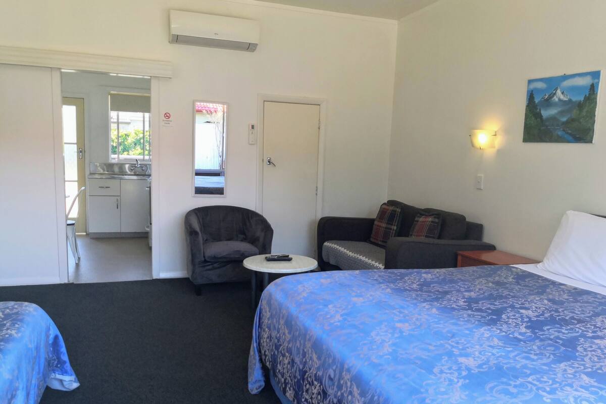 A hotel room with two beds, a sofa, a chair, a small table, a painting, and a kitchenette.