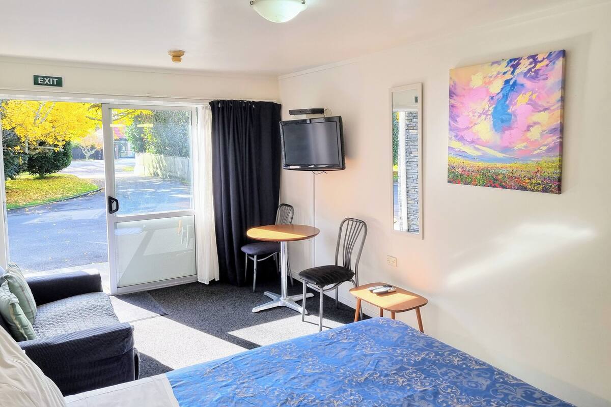 A hotel room with a bed, a small table, a chair, a TV, and a painting on the wall.